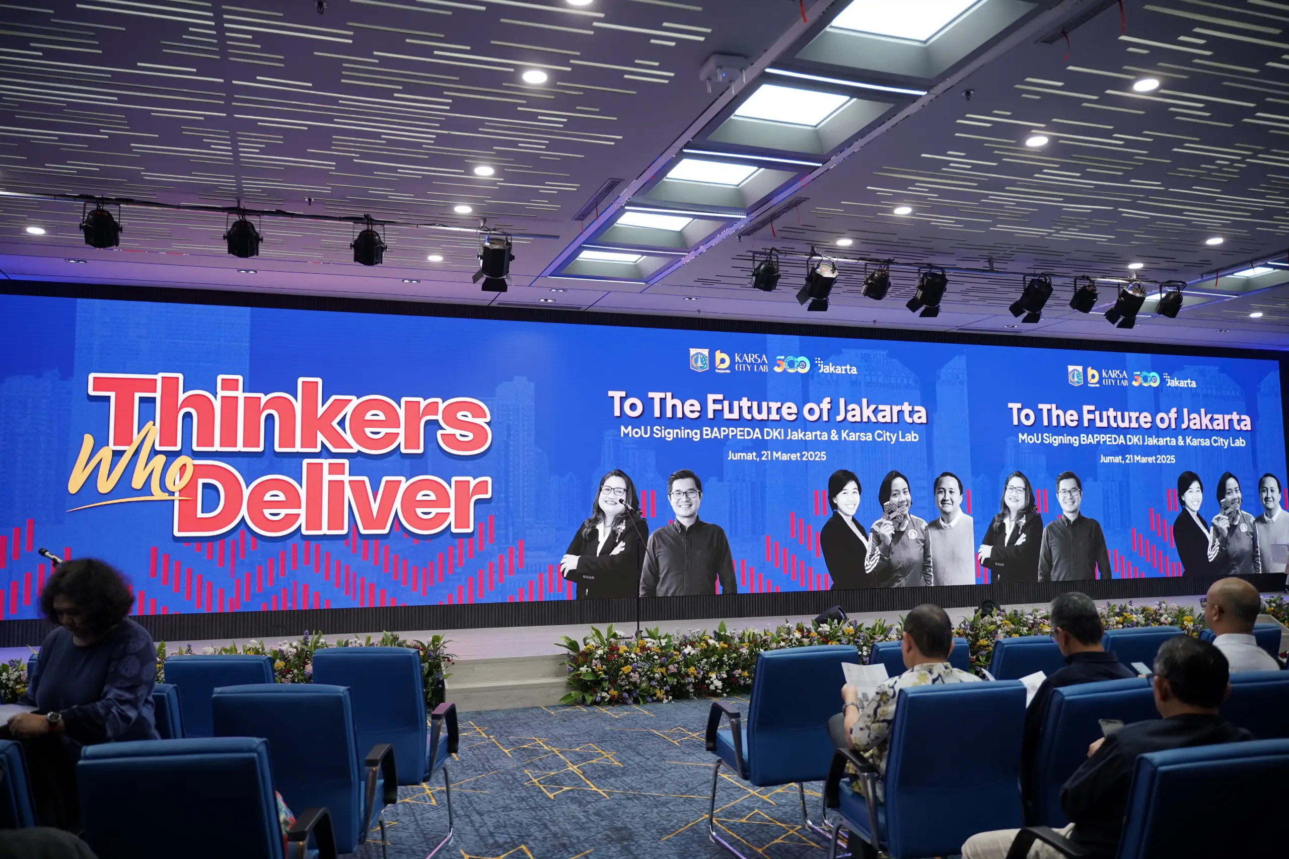 A conference room with blue chairs, floral arrangements, and a large screen displaying "Thinkers Who Deliver" and an MoU signing event.